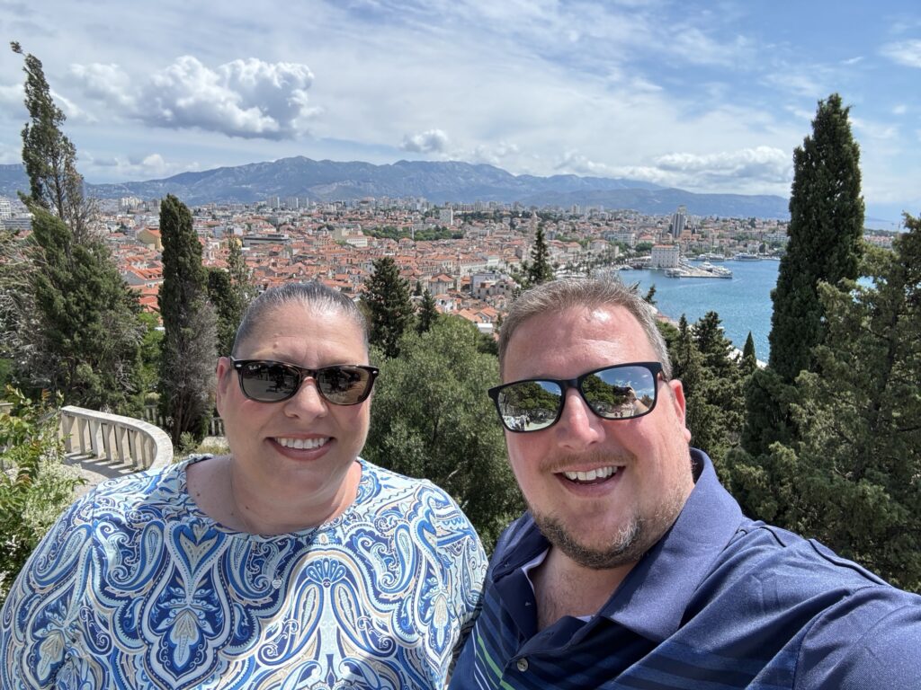 A smiling woman and man wearing sunglasses take a selfie outdoors with a scenic city, coastline, and mountains in the background, surrounded by tall trees under a partly cloudy sky.