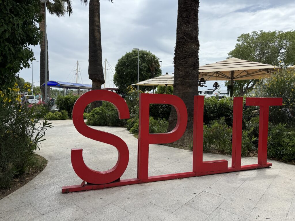 Large red letters spelling SPLIT stand on a paved walkway, surrounded by palm trees and greenery. Outdoor seating with umbrellas and a marina with boats are visible in the background under a cloudy sky.