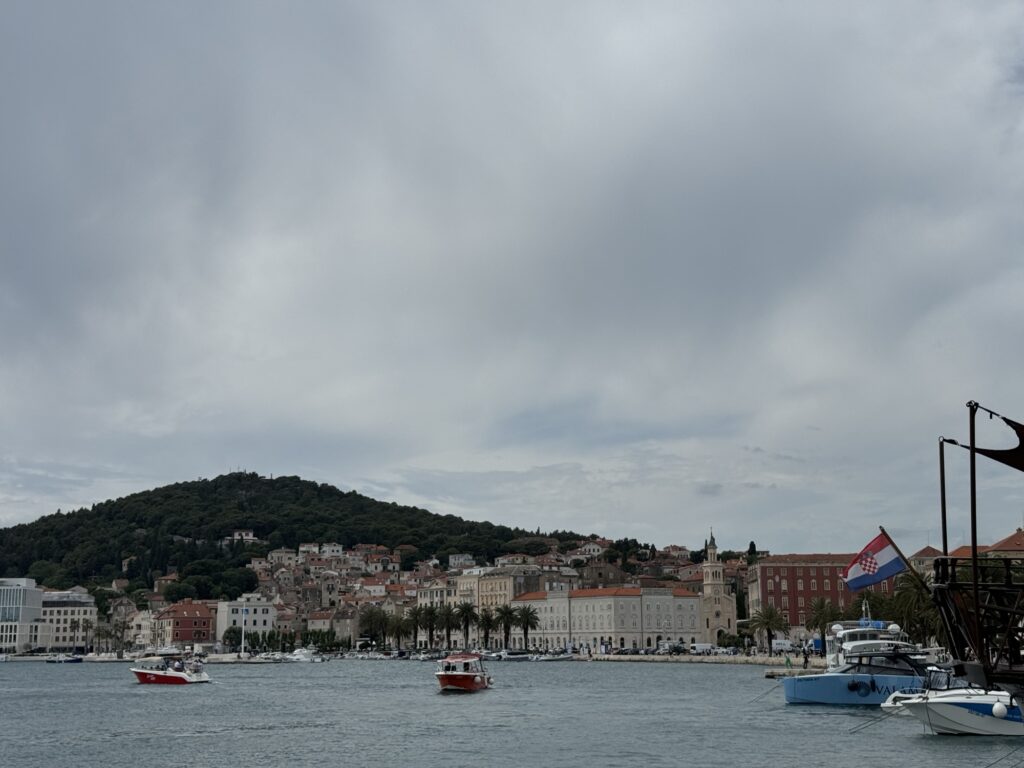 A view of a coastal town with red-roofed buildings and a hill in the background. Boats are anchored and moving on the water, and a Croatian flag is visible on a docked boat to the right under a cloudy sky.