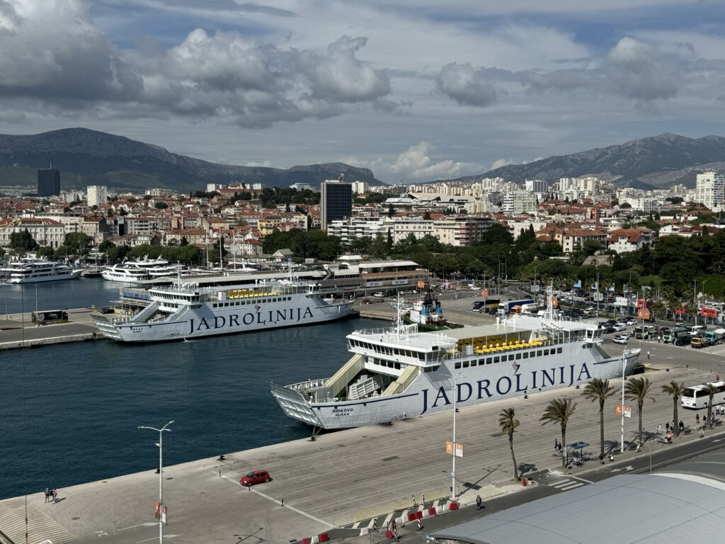 Two large Jadrolinija ferries are docked at a harbor in Split, Croatia, with city buildings and mountains visible in the background under a partly cloudy sky.