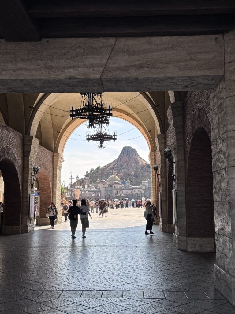 View through arched stone entrance at Tokyo DisneySea with Mount Prometheus rising in the background and visitors walking toward Mediterranean Harbor