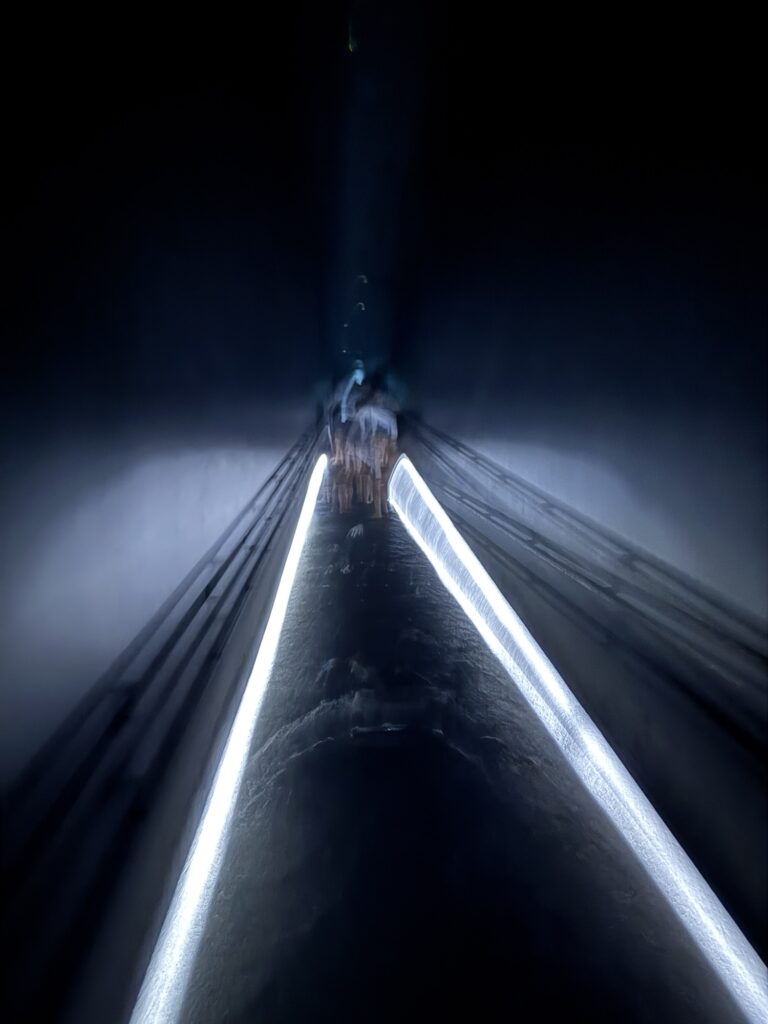 A dark, abstract image showing a reflective surface with two parallel lines of bright white light converging toward a vanishing point, creating a tunnel-like effect.