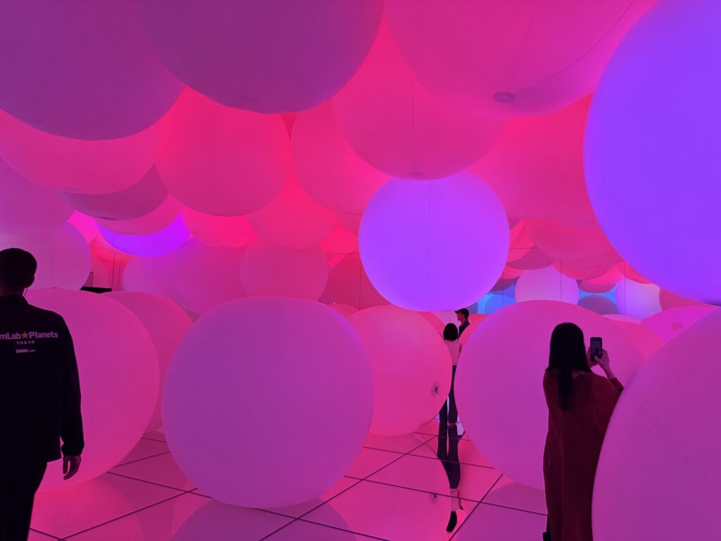 People stand among large, glowing white spheres in a room bathed in pink and purple light. One person takes a photo, and the reflective floor enhances the colorful, immersive atmosphere.