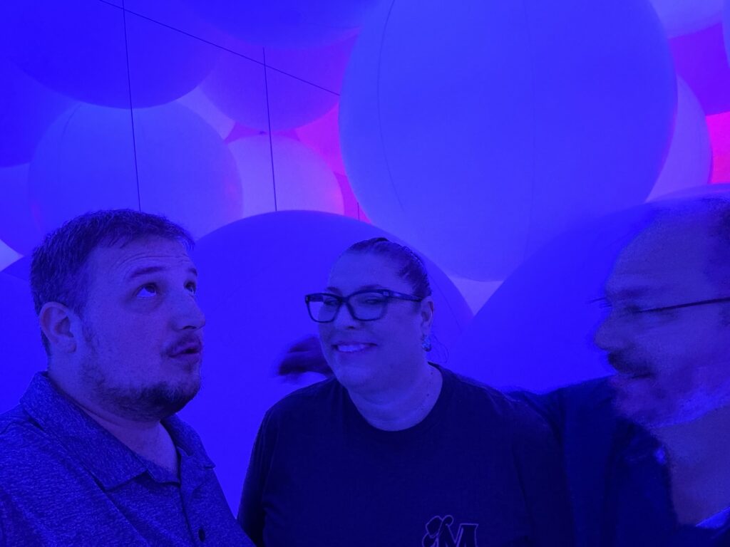 Three adults stand together in a room filled with large, round, balloon-like objects, illuminated by blue and purple lighting. One person looks up, another smiles, and the third appears slightly blurred in motion.