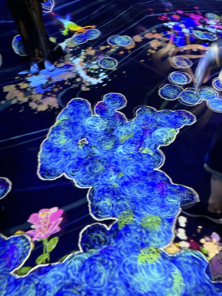 A vibrant digital floor display shows swirling blue shapes, colorful splashes, and flowers. Several peoples legs and feet are visible standing on the illuminated artwork.