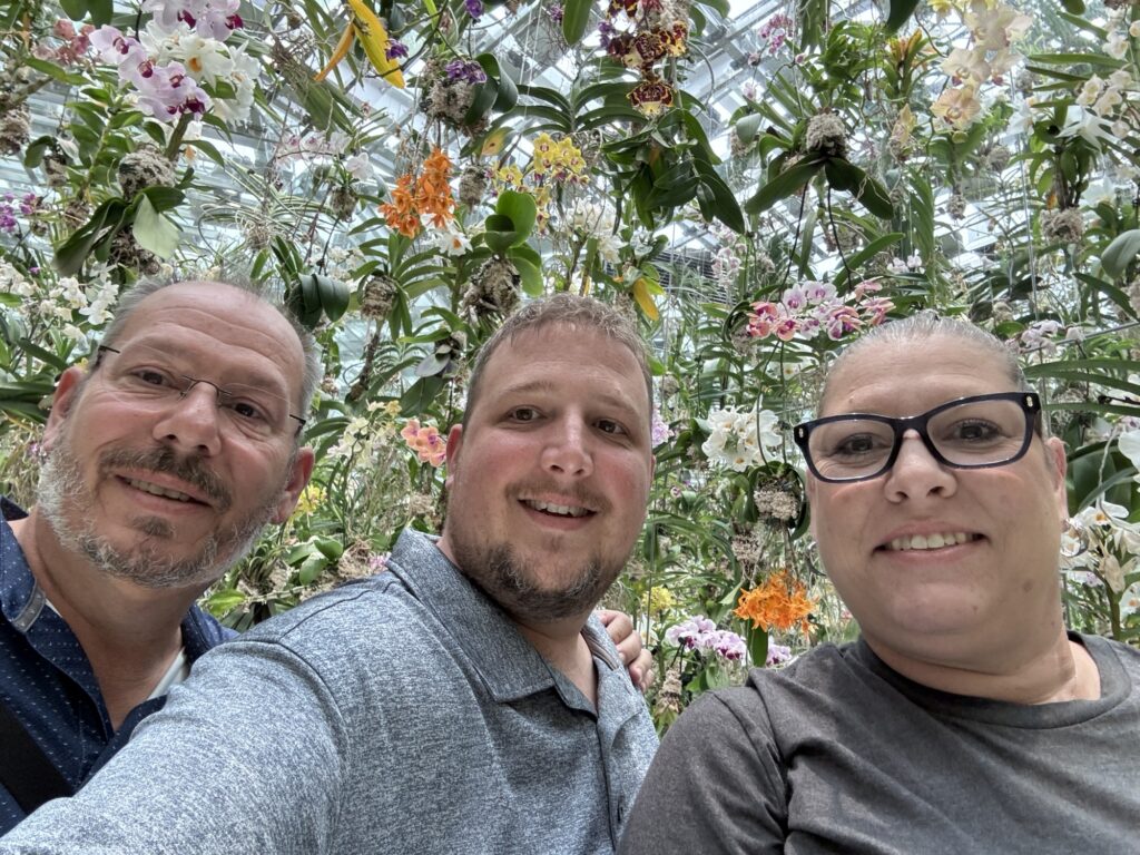 Three people pose for a selfie in front of a lush display of colorful orchids and greenery in a greenhouse, smiling at the camera with flowers and leaves filling the background.
