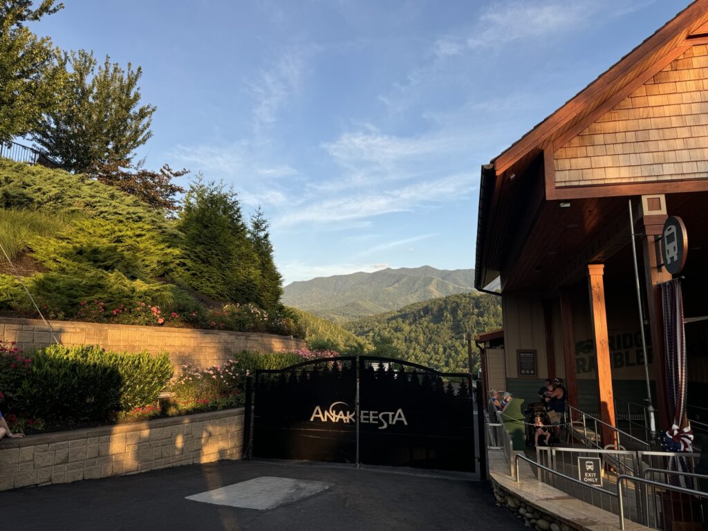 Entrance gate to Anakeesta with mountains and lush greenery in the background, a wooden building on the right, and people near the entrance under a clear blue sky at sunset.