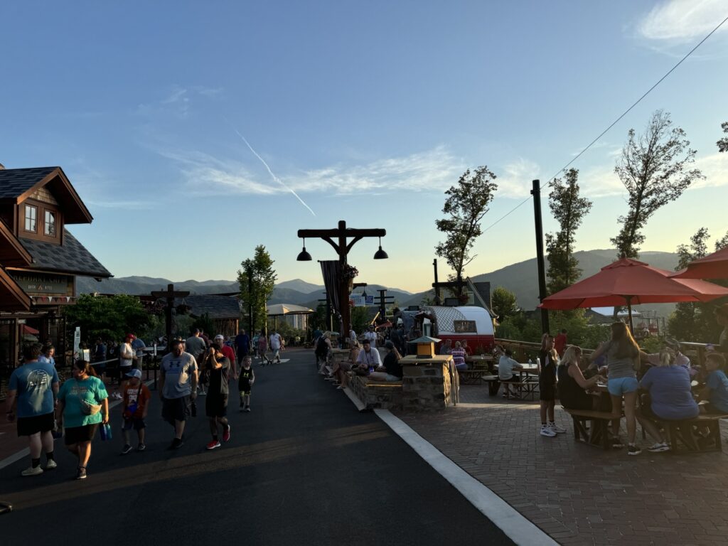 People stroll along a paved path and sit at outdoor tables under red umbrellas at a lively park or resort in the evening, with mountains and blue sky in the background.