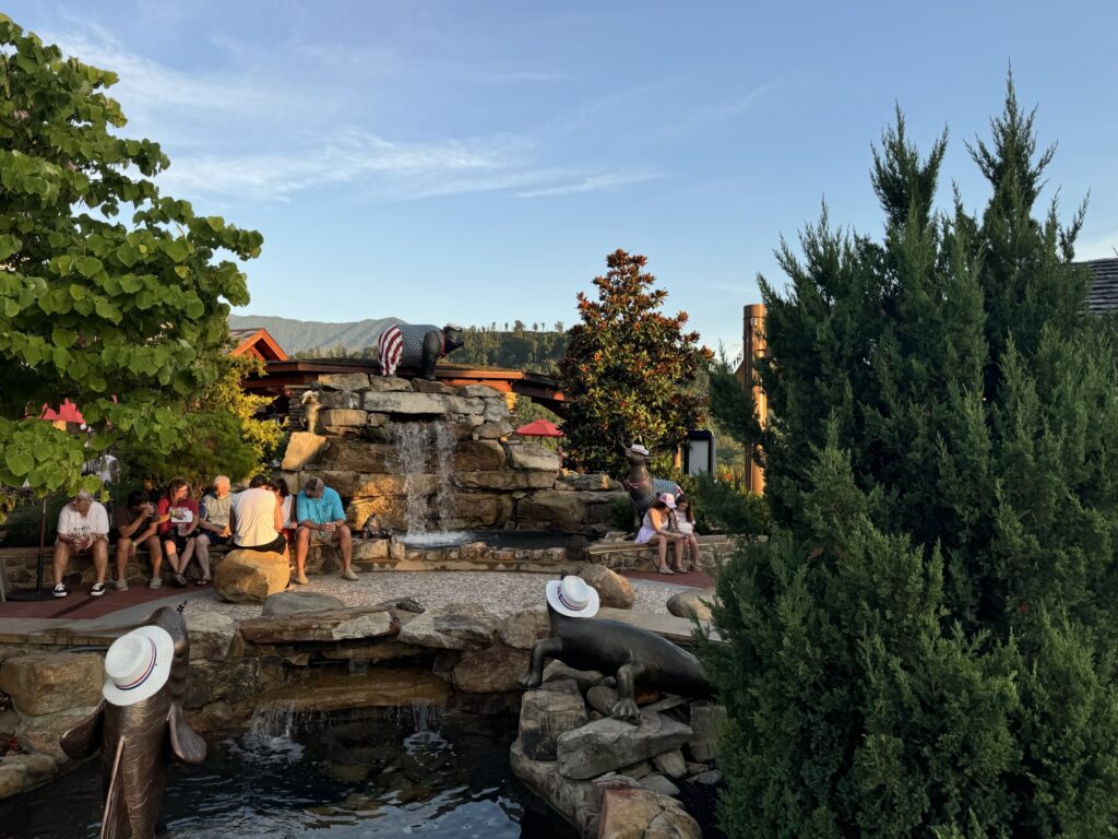 People sit and relax around a stone waterfall feature in a landscaped outdoor area with trees, greenery, and dolphin statues by a pond, under a clear blue sky at sunset.