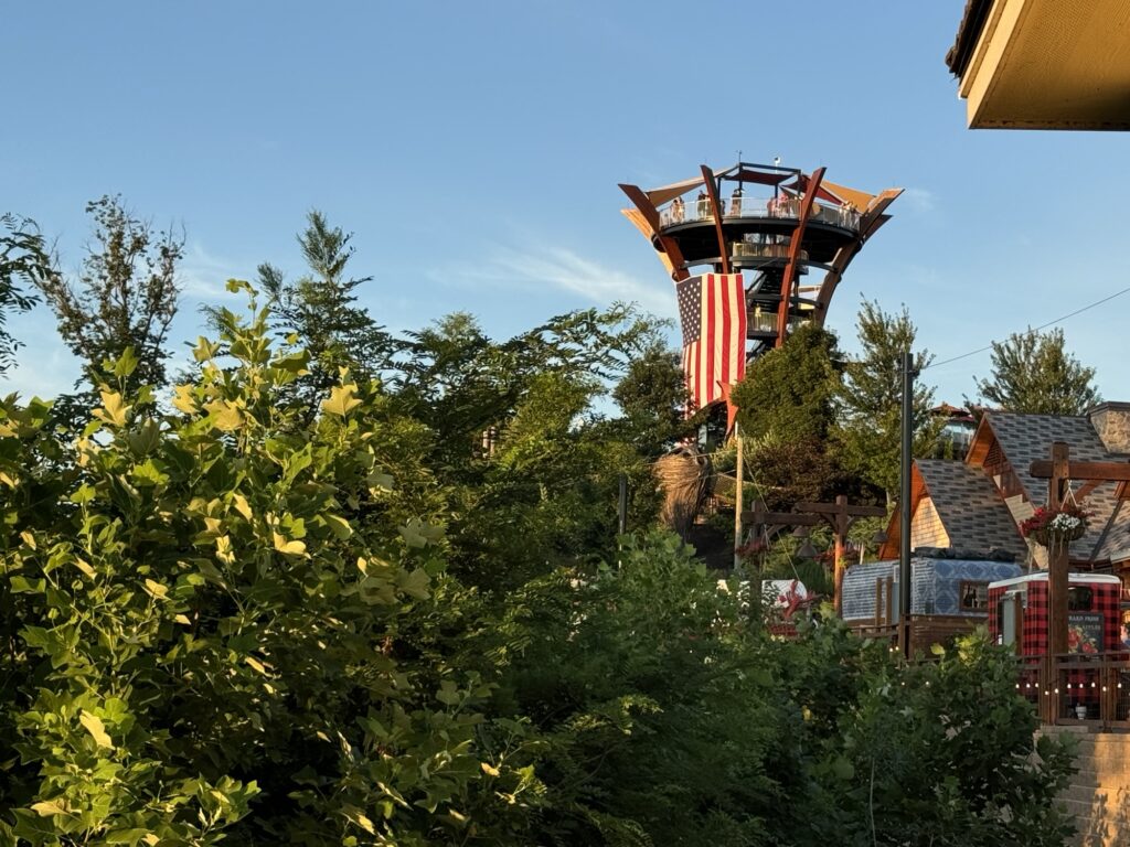 A tall, unique wooden tower rises above green trees and buildings. An American flag hangs prominently in the center, and the sky is clear and blue, suggesting a sunny day.