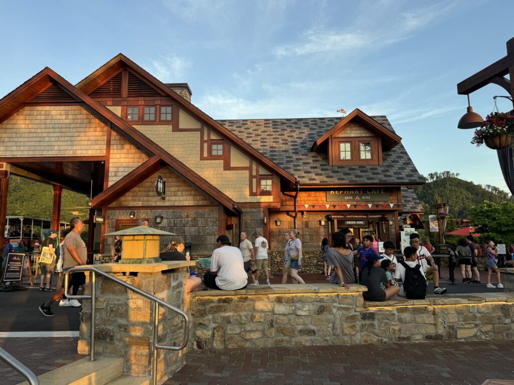 People gather outside a rustic-style café and market building with wood and stone exterior at sunset. Some sit on a stone wall, while others stand or walk nearby. Flower pots and hills are visible in the background.