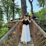 Three people smile while standing on a rope bridge surrounded by trees. A woman in a white dress stands in front, with two men standing behind her. The bridge leads to a wooden platform among the trees.