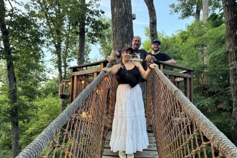 Three people smile while standing on a rope bridge surrounded by trees. A woman in a white dress stands in front, with two men standing behind her. The bridge leads to a wooden platform among the trees.