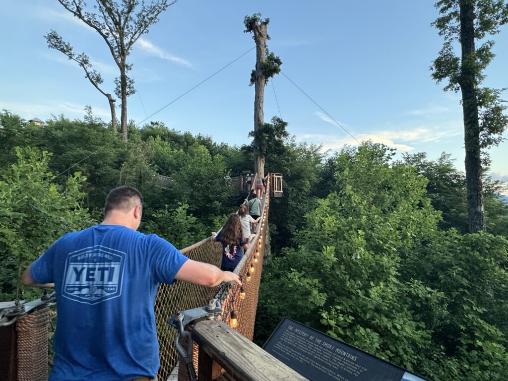 People walk on a rope bridge surrounded by lush green trees, with a man in a blue “YETI” shirt in the foreground. The bridge is elevated and stretches towards a wooden platform ahead.