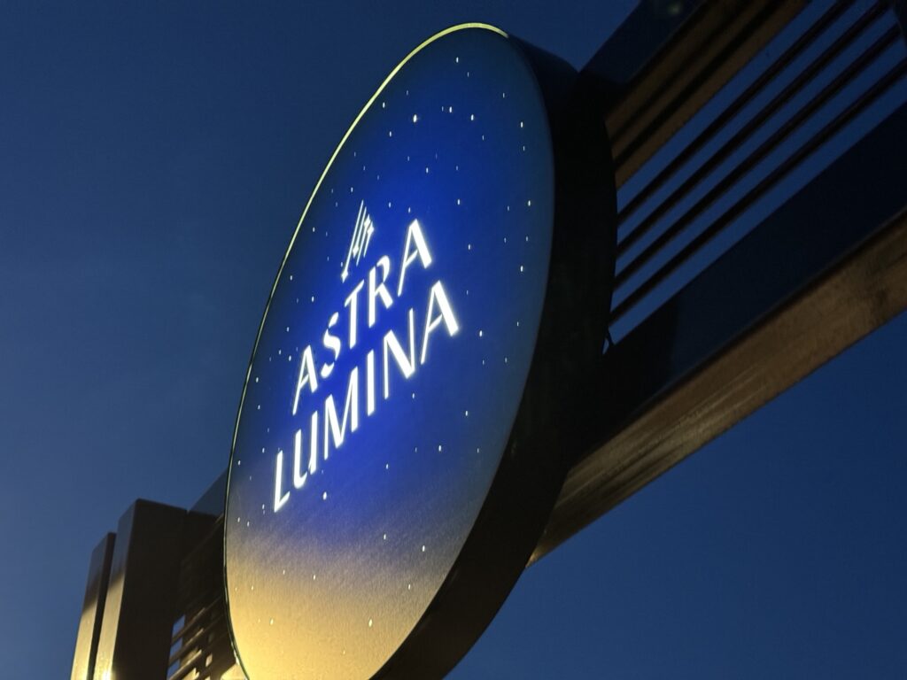 A round, illuminated sign with a starry background displays the words ASTRA LUMINA in white letters, mounted on a metal structure against a dark blue evening sky.