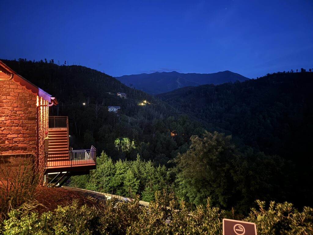 A warmly lit cabin sits on a hillside overlooking a lush, tree-covered valley at dusk, with distant houses and mountain silhouettes visible under a deep blue sky.