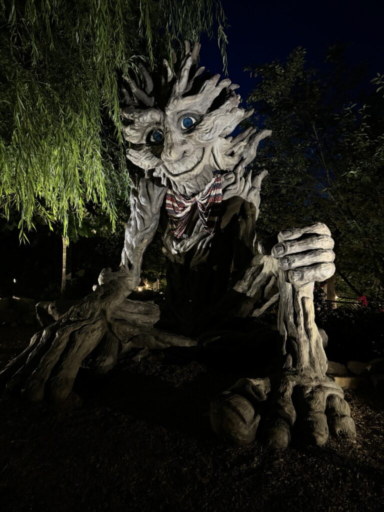 A large tree-like sculpture with a smiling face, blue eyes, and a red-and-white scarf sits outdoors at night, illuminated by spotlights and surrounded by foliage.