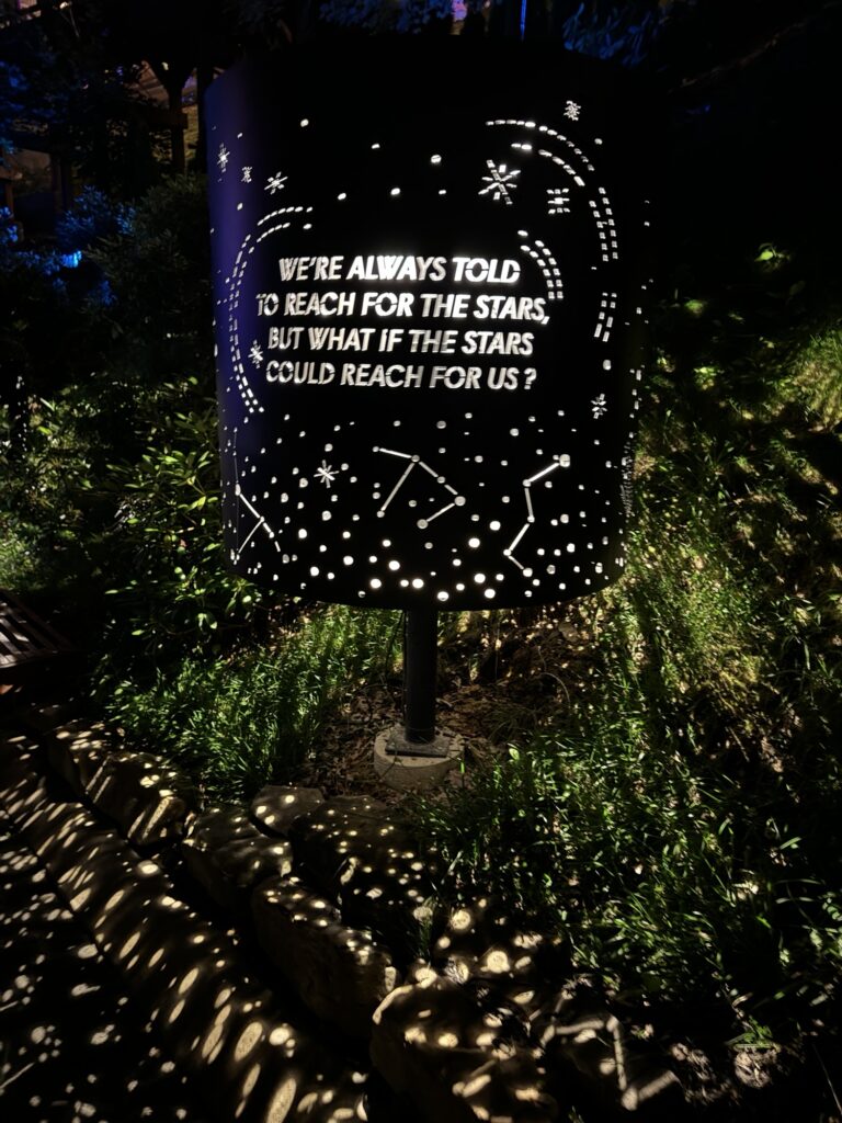 A cylindrical sign glows at night, featuring constellations and star patterns, with the words: Were always told to reach for the stars, but what if the stars could reach for us?.