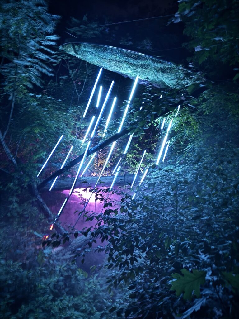 Blue vertical light tubes hang among dense green foliage, illuminating the leaves with a neon glow. A purple light shines in the background, creating a dreamy, surreal forest scene at night.