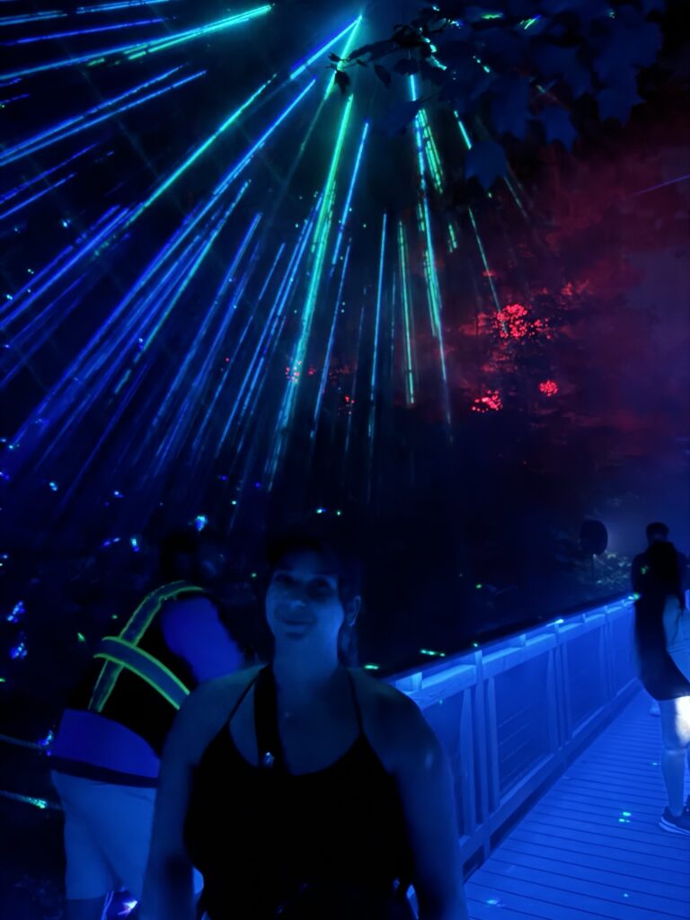 A person stands on a lit pathway at night, surrounded by vibrant blue and green laser lights beaming through trees. Another person in a reflective vest is further down the path, with a colorful, atmospheric glow in the background.