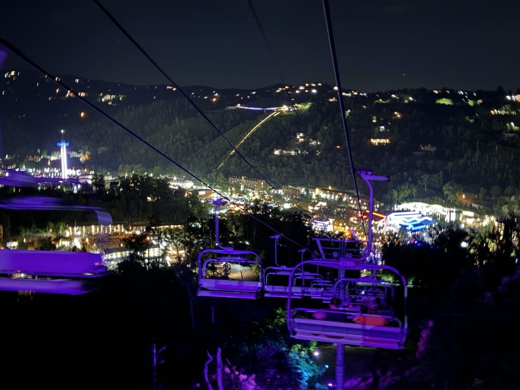 A brightly lit town at night seen from a cable car ride above, with glowing lights from buildings and streets scattered across the hills in the distance. The cable car seats and cables are visible in the foreground.