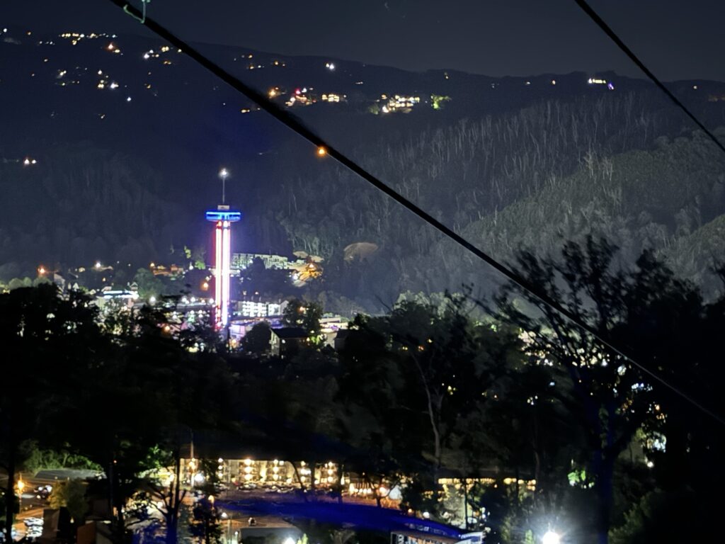 A nighttime cityscape with a brightly lit observation tower in the center, colorful lights shining, surrounded by dark hills and trees, with distant houses visible on the hillside.