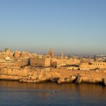 Golden-hour view of Valletta, Malta, with historic sandstone buildings and domed churches glowing in warm light above fortified walls along the waterfront, as a small boat moves across calm blue water.