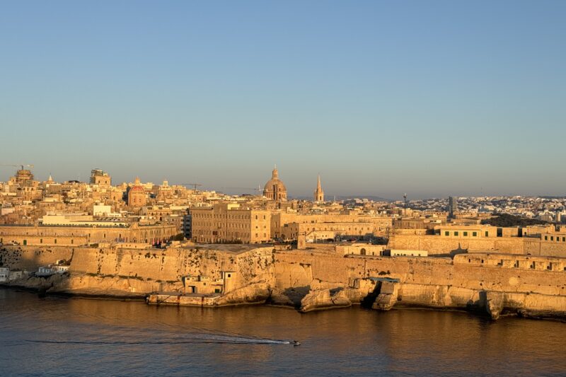 Golden-hour view of Valletta, Malta, with historic sandstone buildings and domed churches glowing in warm light above fortified walls along the waterfront, as a small boat moves across calm blue water.