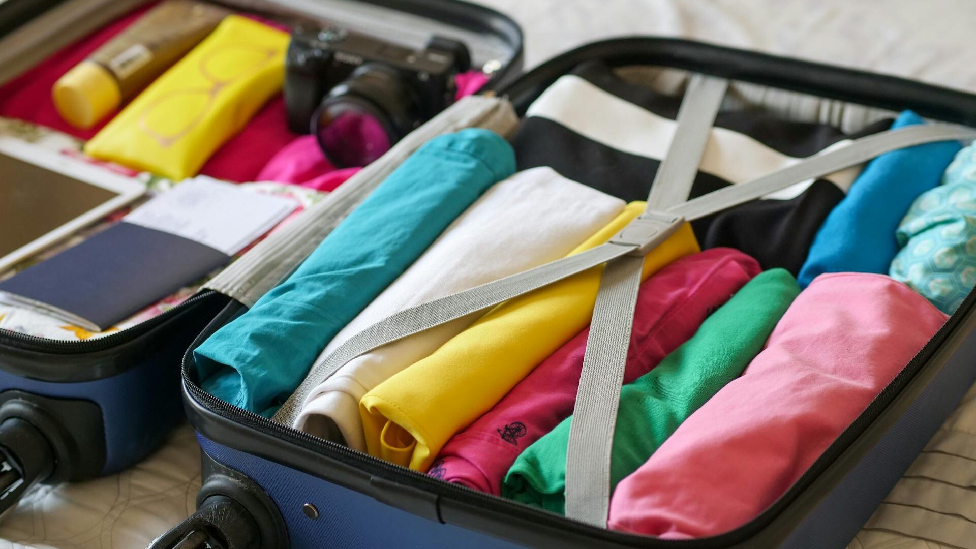 Image of a blue clamshell suitcase with rollers, laying on a bed, filled with colorful rolled clothes on the right, and a passport, camera, and sunscreen on left left side.