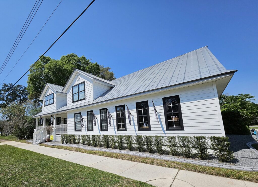 Modern white farmhouse with metal roof and black-trimmed windows on a sunny residential street with green lawn