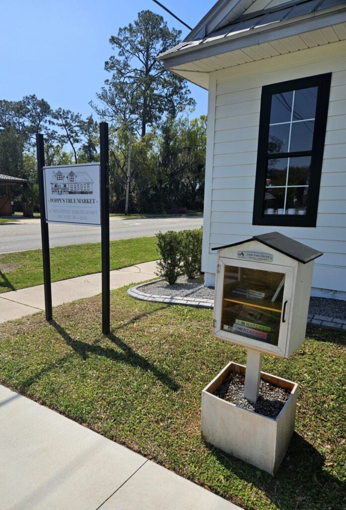 A little free library stands in front of Poppy's Tree Market, a white building with black-trimmed windows and a posted sign