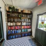 A well-organized library room with floor-to-ceiling bookshelves organized by color, featuring plants, patterned flooring, and an exit sign