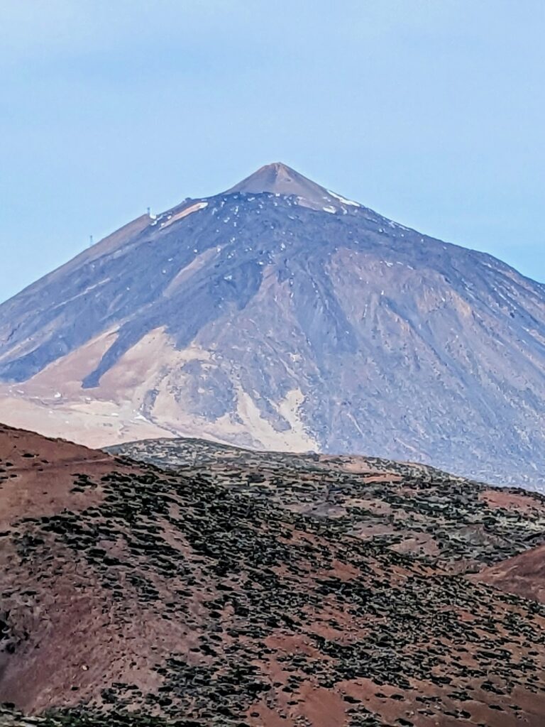 A tall, conical mountain with a rocky, barren slope and patches of snow at the summit, set against a blue sky with rugged, reddish-brown terrain in the foreground.
