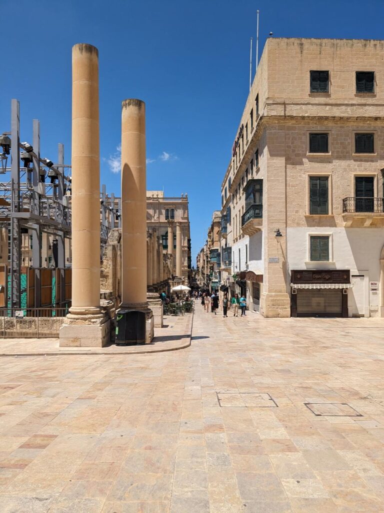 Open stone plaza with tall columns and historic buildings, leading to a narrow street with pedestrians under a clear blue sky.