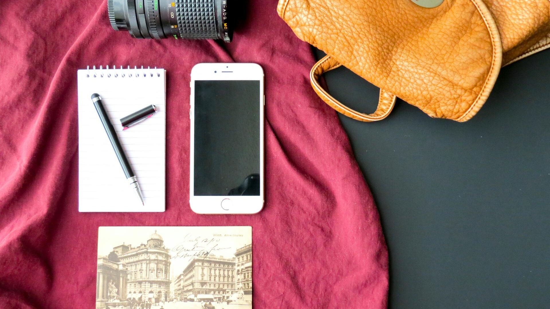A flat lay of travel essentials on a red cloth: a camera lens, brown leather bag, notepad with a pen, smartphone, and a vintage postcard with an old city scene.