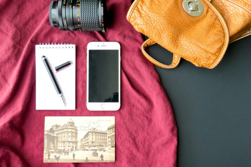 A flat lay of travel essentials on a red cloth: a camera lens, brown leather bag, notepad with a pen, smartphone, and a vintage postcard with an old city scene.