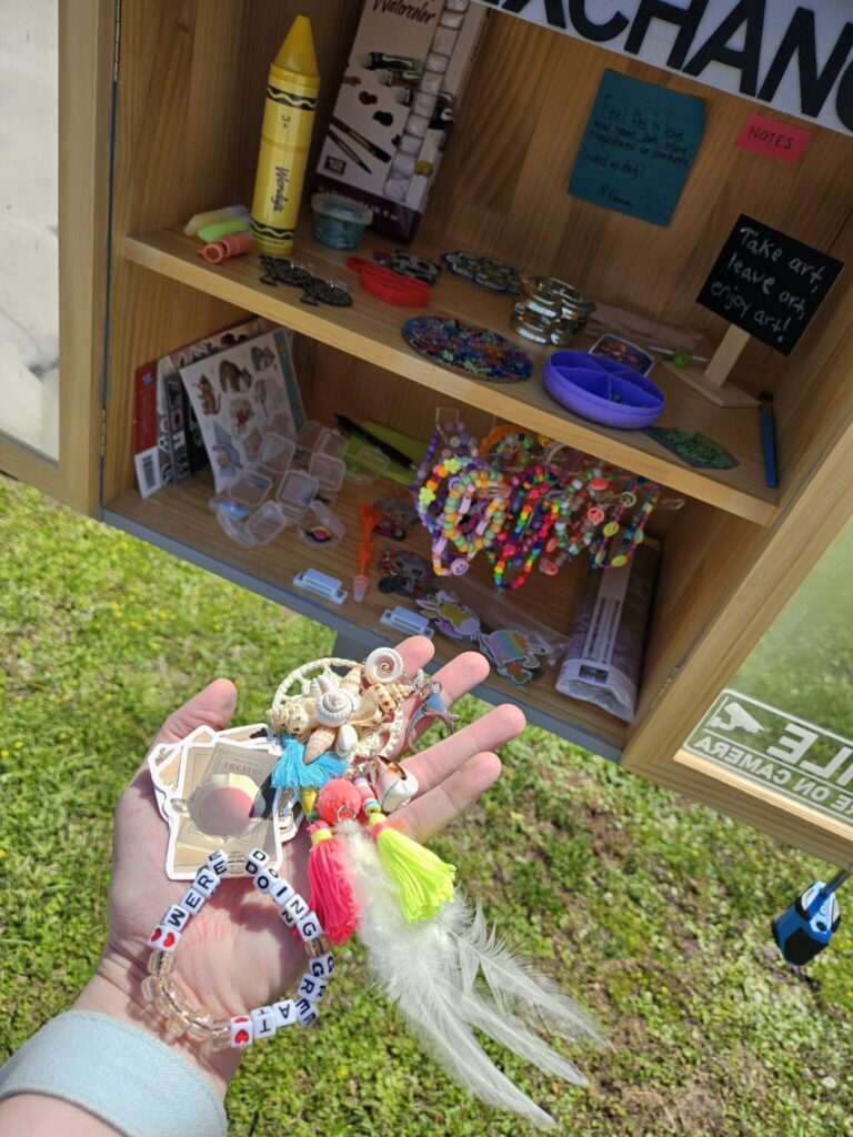 A hand holding a colorful beaded keychain with tassels and feathers, with a wooden shelf display of crafts and accessories in the background