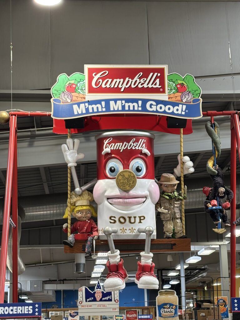 Campbells brand soup can swinging on a swing with the campbells boy and girl. Large display hanging from the ceiling. There is also two monkeys hanging down next to the swing. This is keeping with the jungle theme at the store.