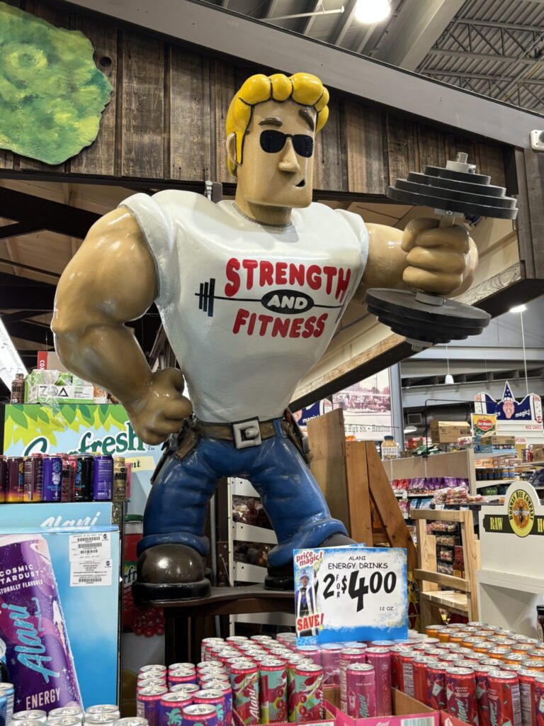 Cartoon like body builder holding a barbell with a display of Alani brand drinks. The sign is showing two for $4.00 at Jungle Jim's grocery store.