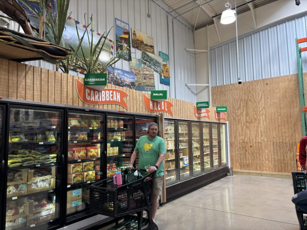 Man pushing a cart in front of freezers filled with frozen food. Signs showing the caribbean, Halal, Spanish/Portuguese and kosher items.