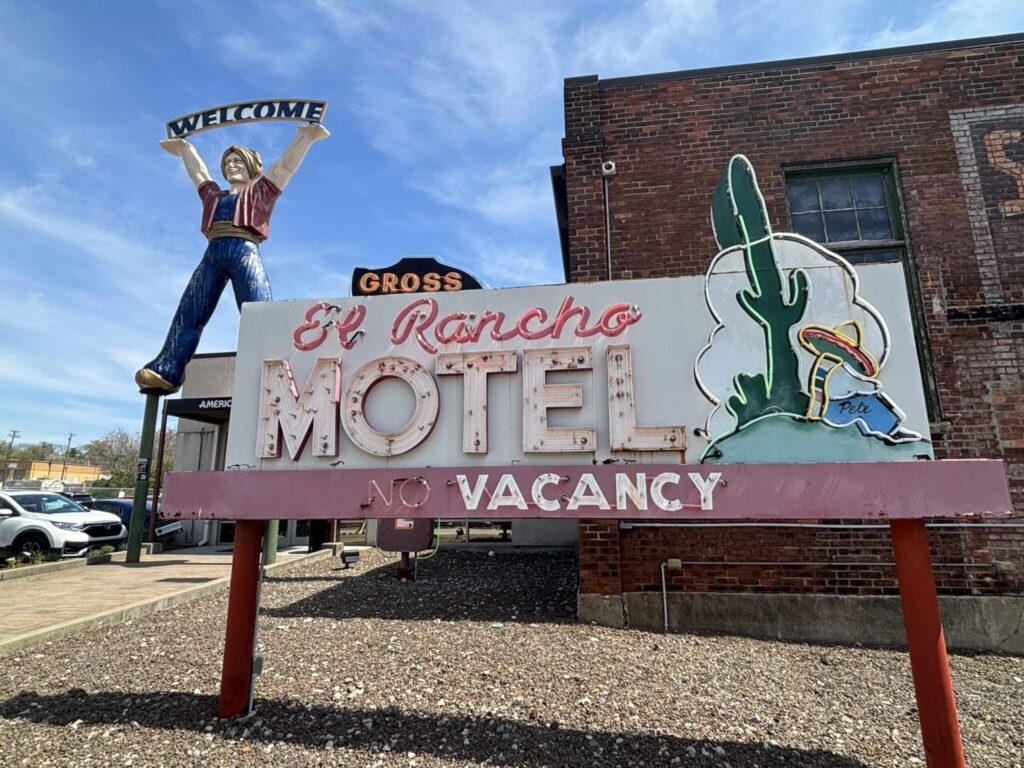 Neon signs in a gravel lot for El Rancho Motel with No vacancy. In the background is a replica of a large woman holding a welcome sign above her head. 