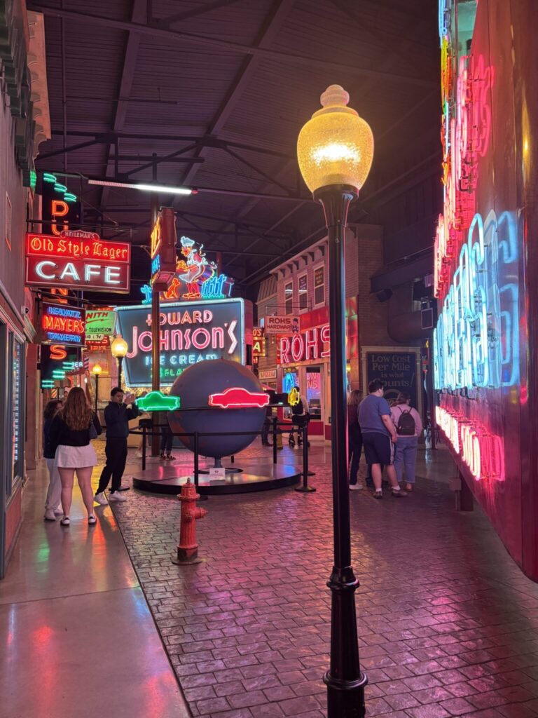 Neon-lit vintage street scene at night with retro signs, street lamp, and visitors walking through an indoor arcade or covered alley
