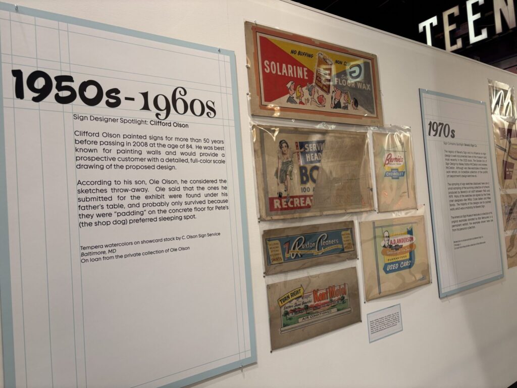 Wall exhibit about 1950s–1960s sign design, featuring a large informational panel about sign painter Clifford Olson alongside several vintage illustrated advertising signs.