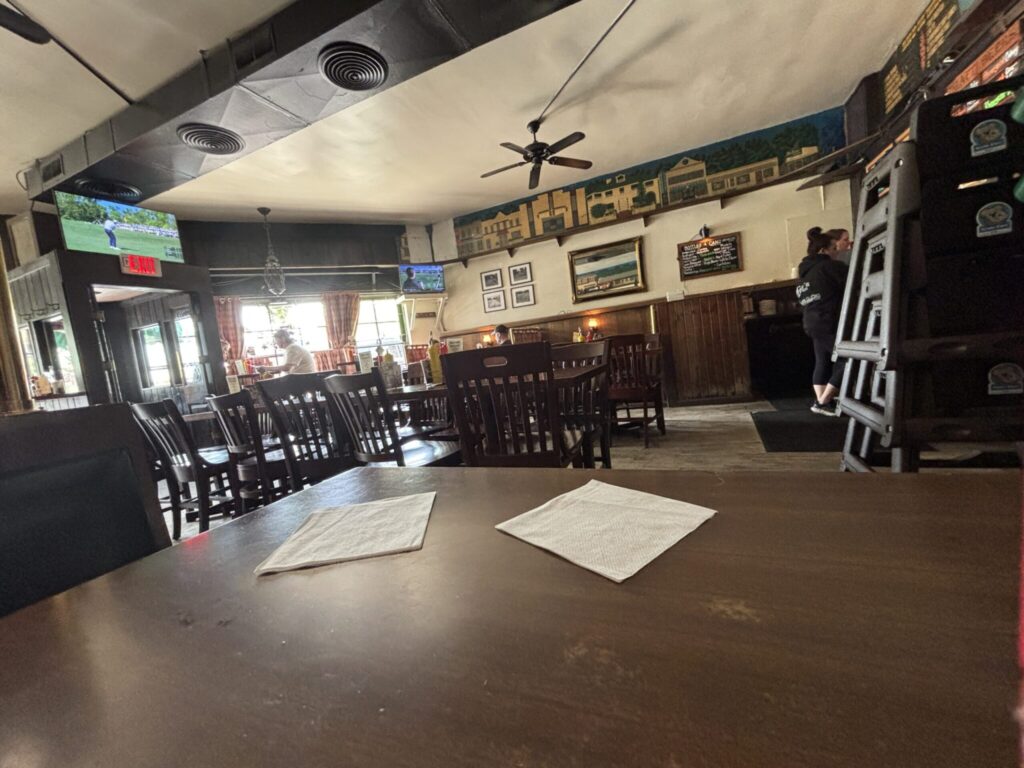 Cozy tavern-style dining room with wooden tables and chairs, a bar area, wall decor, and televisions showing sports, with a few people seated and natural light coming through front windows.