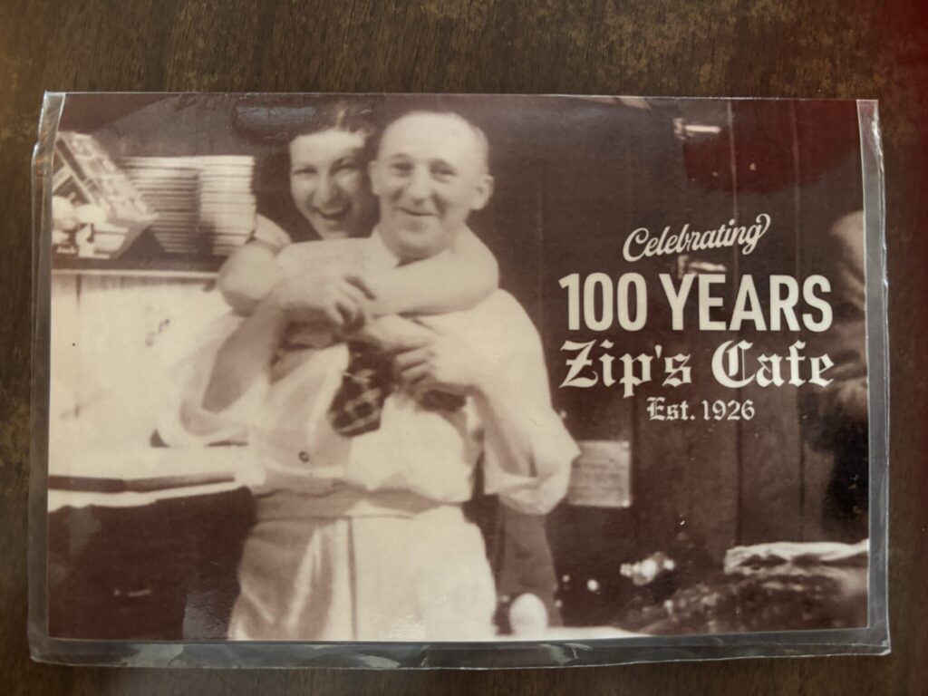 Sepia-toned vintage photo of a smiling man and woman in a kitchen setting, displayed with text reading 'Celebrating 100 Years Zip's Cafe Est. 1926,' highlighting the restaurant's long history."