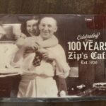 Sepia-toned vintage photo of a smiling man and woman in a kitchen setting, displayed with text reading 'Celebrating 100 Years Zip's Cafe Est. 1926,' highlighting the restaurant's long history."
