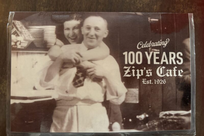 Sepia-toned vintage photo of a smiling man and woman in a kitchen setting, displayed with text reading 'Celebrating 100 Years Zip's Cafe Est. 1926,' highlighting the restaurant's long history."