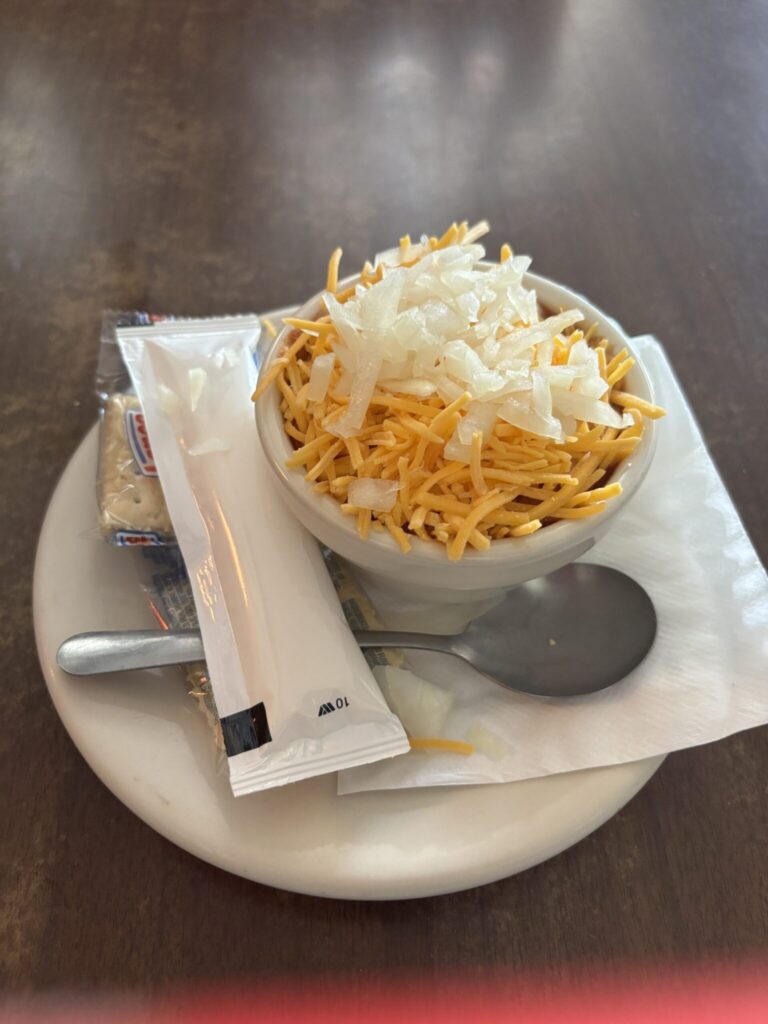 A cup of chili topped with onion and cheese. On the saucer is a napkin, spoon, crackers and a pack of sour cream.