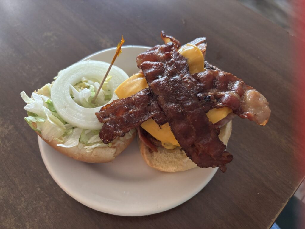 A Zips's Cafe burger topped with ham, bacon and american cheese. The top bun is topped with lettuce and onion, help on with a yellow toothpick.