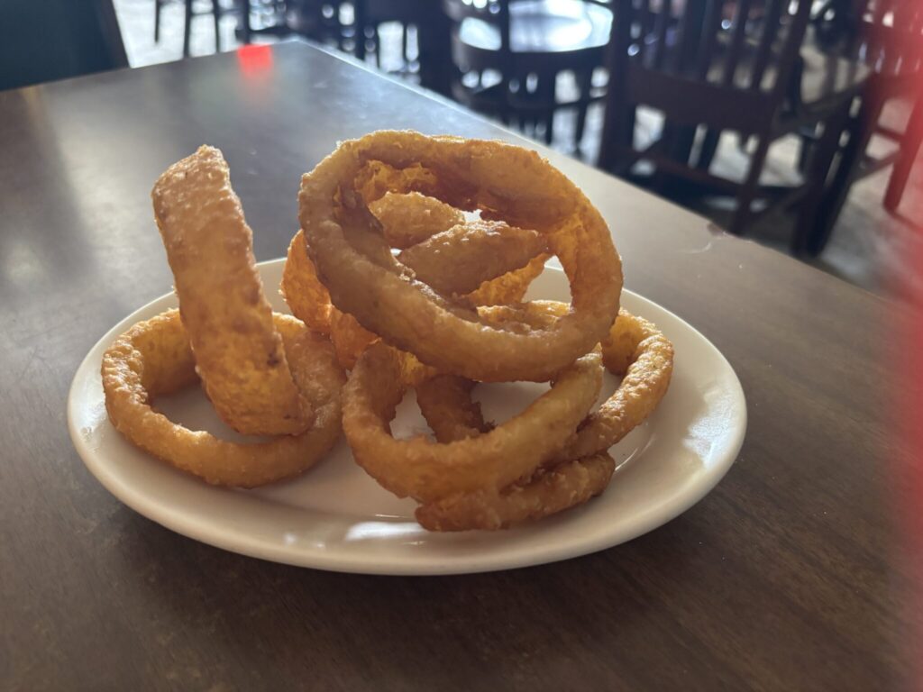 Onion rings piled up on a white oval plate, sitting on a brown table with chairs int he background.